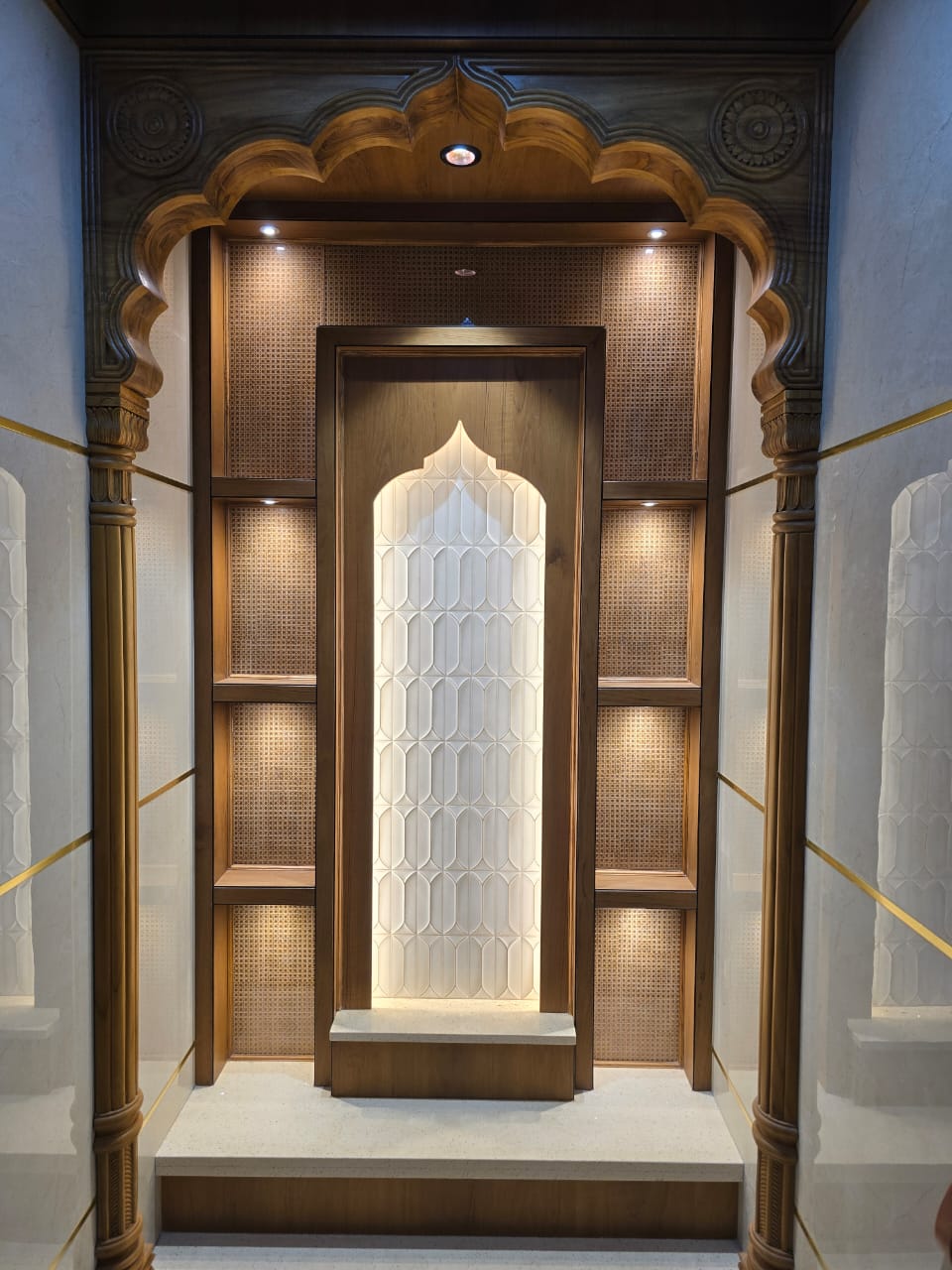 Ornate pooja room with a hand-carved wood jharokha-style arch, a backlit marble jaali screen and cane-panelled side niches.