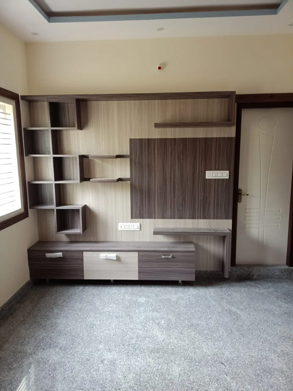 Empty living room with an asymmetric dark-wood cubby shelf and TV panel wall, a stepped cove ceiling, and speckled grey terrazzo flooring.