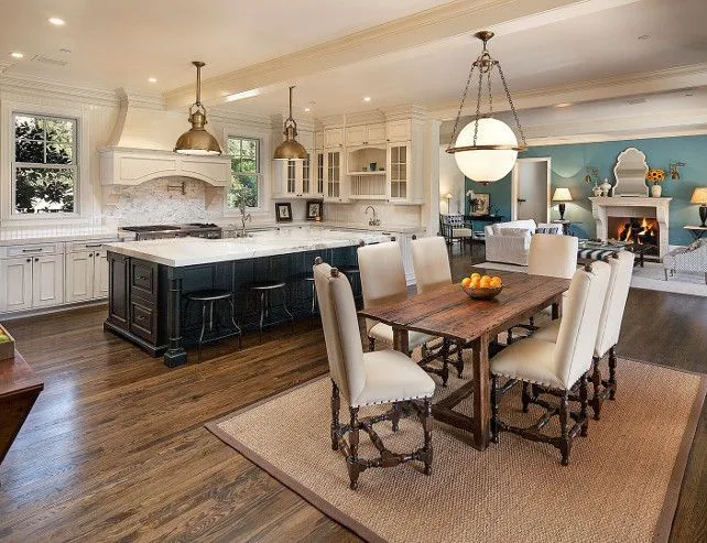 Open-plan kitchen, dining and living area with a dark kitchen island, brass pendant lights, a rustic wood dining table seating eight, and a fireplace beyond.