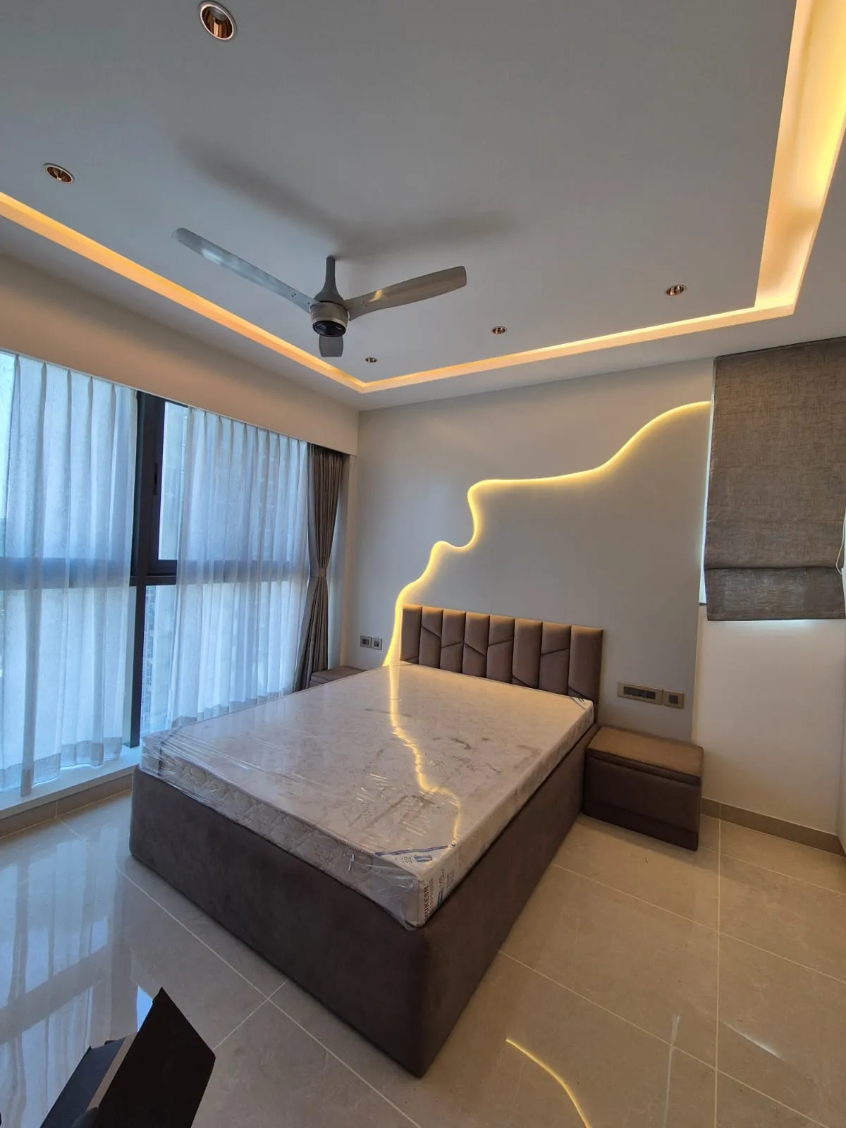 Bedroom photographed at handover with the mattress still shrink-wrapped: taupe upholstered storage bed and matching side stool under a wavy concealed-LED line