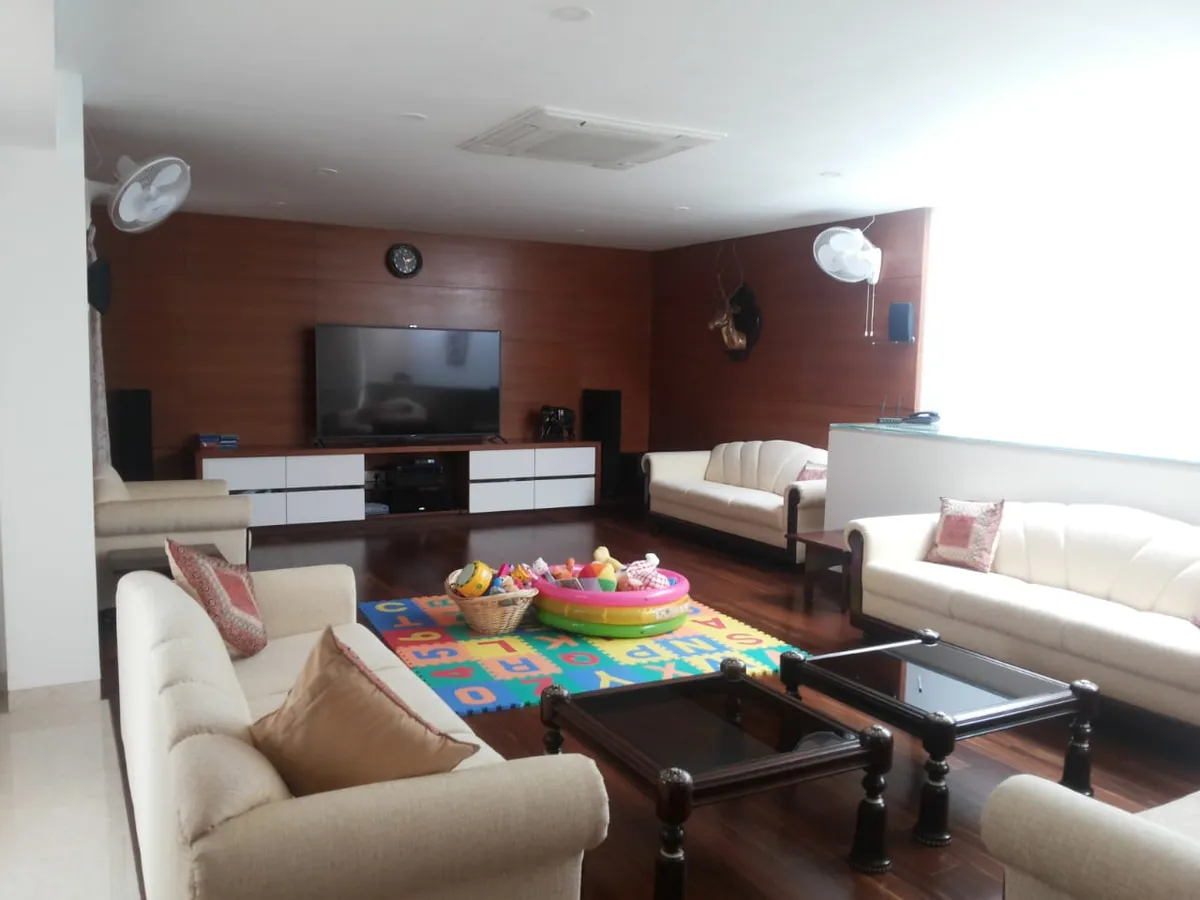 Lived-in family living room with three cream sofas around two carved dark-wood glass-top tables, a teak-panelled TV wall over a white-and-wood console