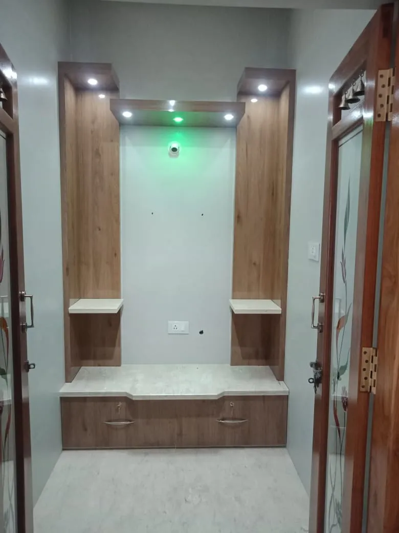 Built-in pooja unit niche with wood-veneer twin towers, a marble seating ledge, storage drawers below and a green accent light illuminating the shrine recess.
