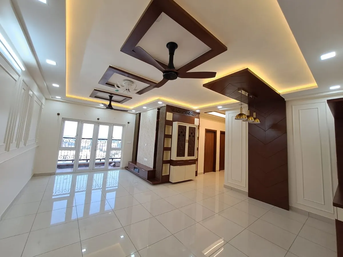 Empty hall with walnut frames set into a cove-lit ceiling over two fans, a carved cream-and-teak pooja cabinet on the far wall