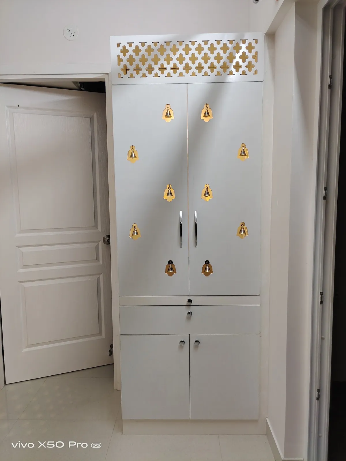 A freestanding white pooja cabinet with a gold cross-pattern jaali vent panel on top and rows of decorative brass bells adorning the closed doors