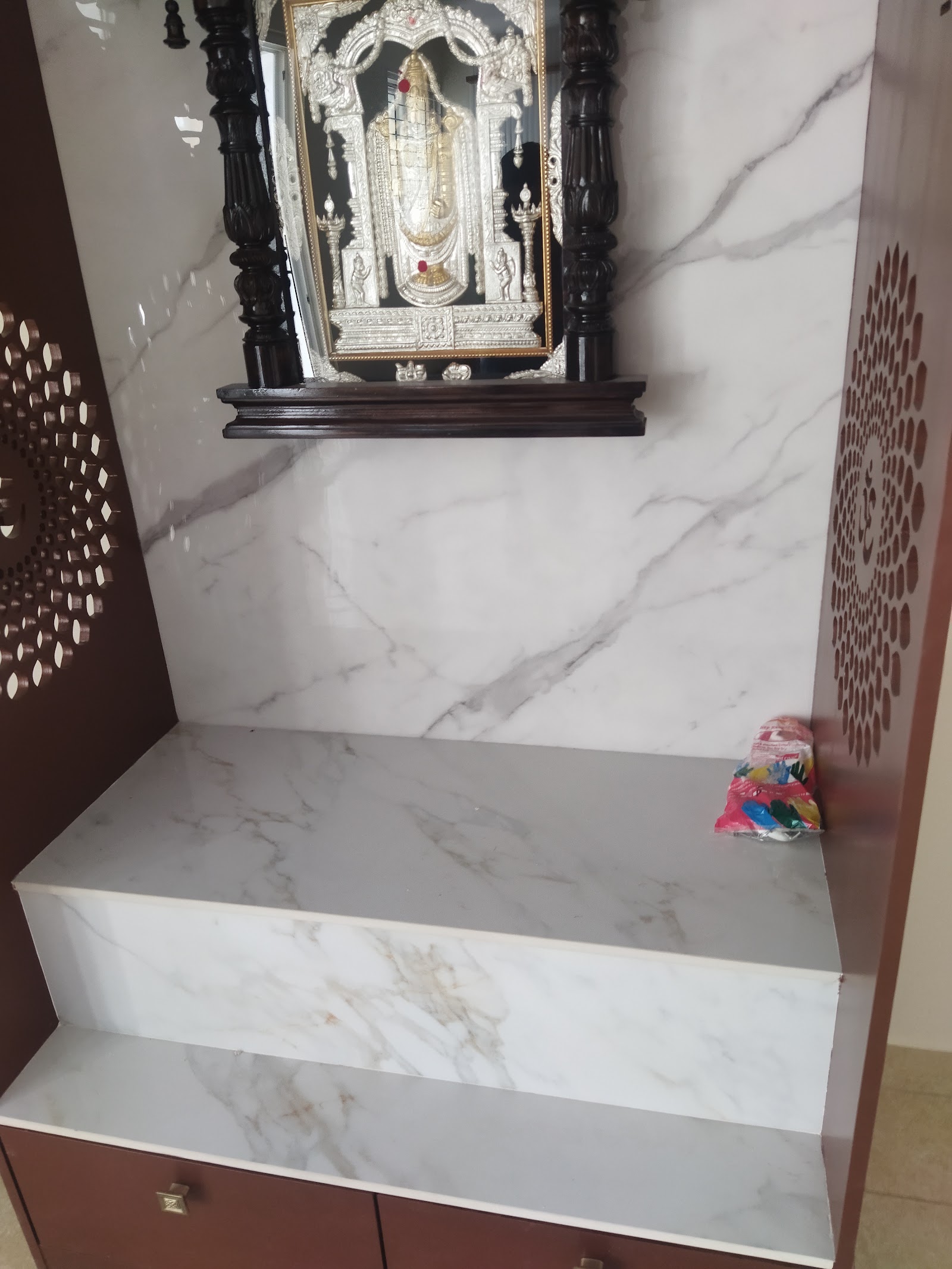Home pooja unit with a marble-clad backwall, a carved dark-wood mandap frame holding a silver Tirupati Balaji idol panel, and jaali side screens.