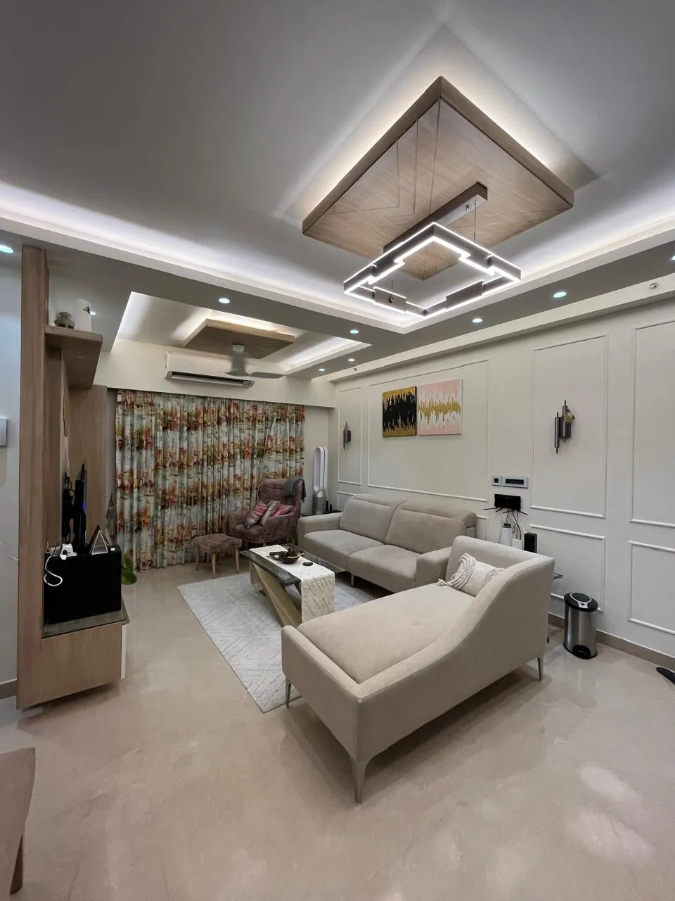 Living room under a wood-clad ceiling raft holding a squared LED profile pendant, with beige fabric sofa and chaise on a cream rug