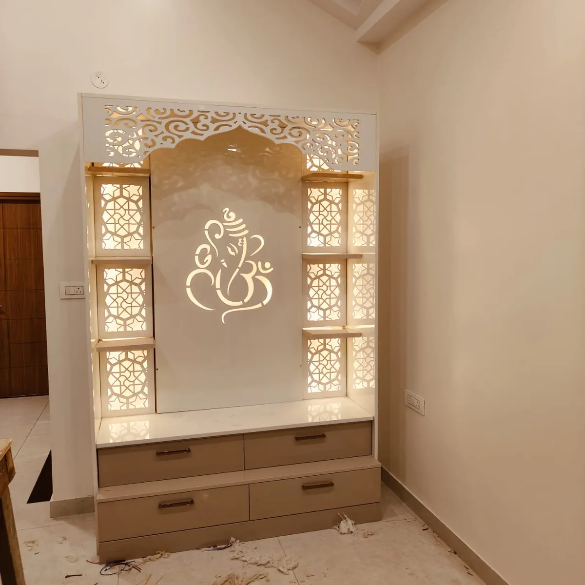 Photograph of a freestanding white-and-beige pooja unit - backlit CNC jaali side panels and a cut-out Ganesha motif over a marble platform