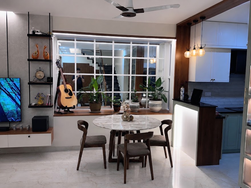 Open-plan dining nook with a round marble-top table and dark wood chairs beneath a large window ledge styled with potted plants