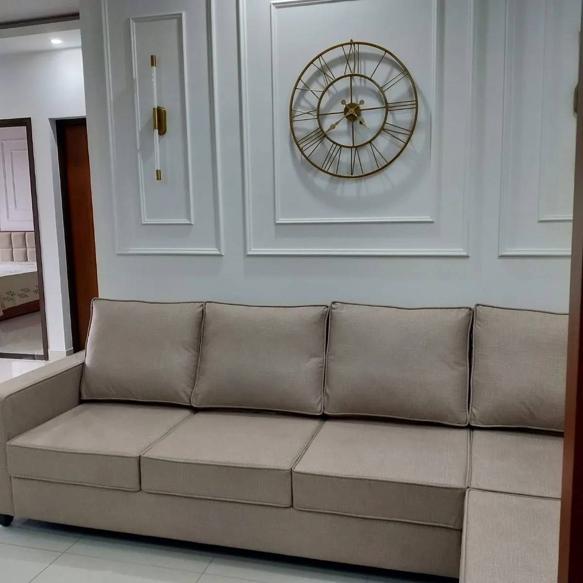 Beige sectional set against a pale blue-grey wall of white PU moulding panels, an oversized gold skeleton clock centred in one panel and a slim brass-and-glass