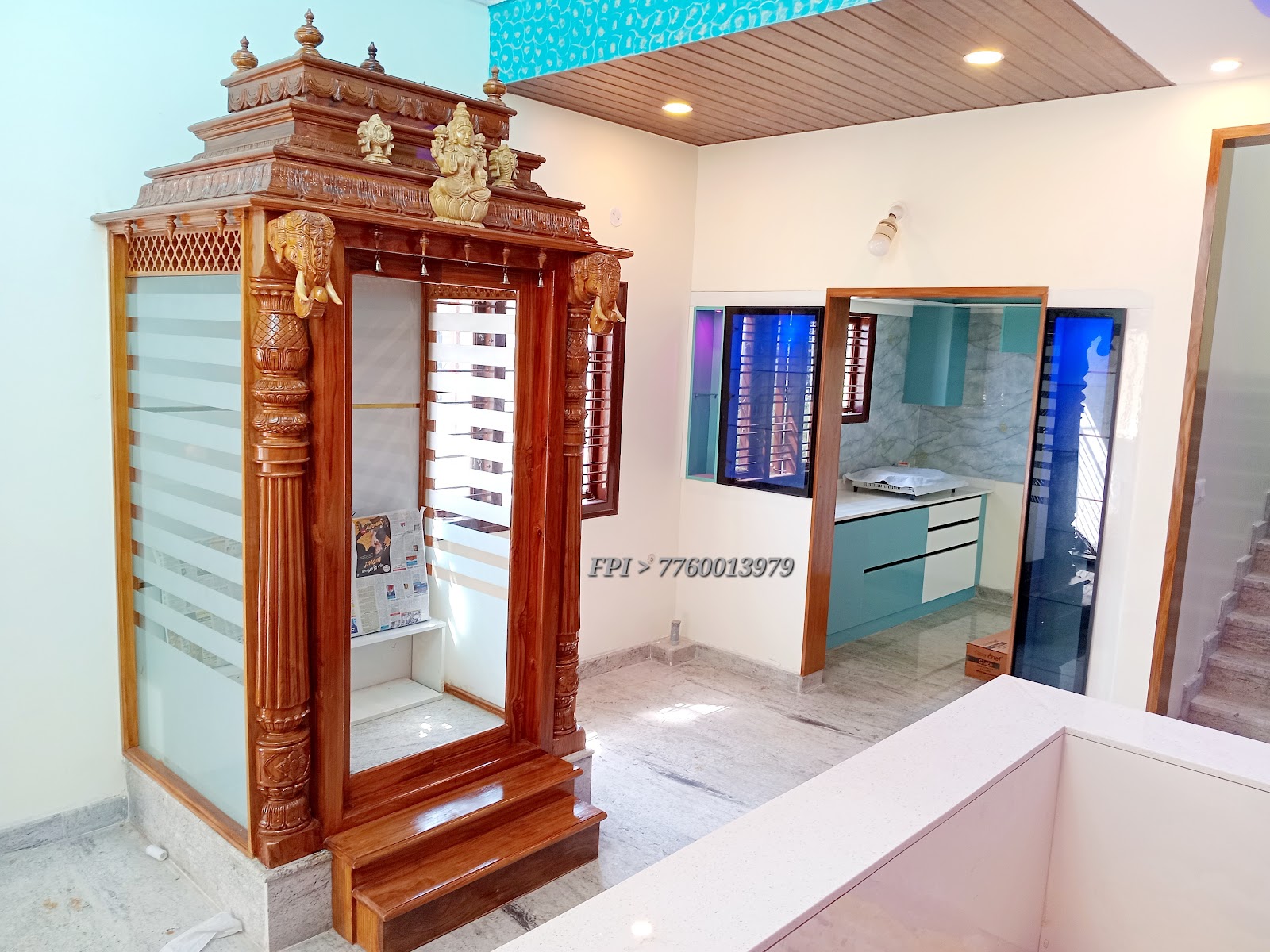 Carved teak pooja mandir with elephant-motif pillars and frosted glass shutters, set in a marble-floored alcove next to an open teal-cabinet kitchen.