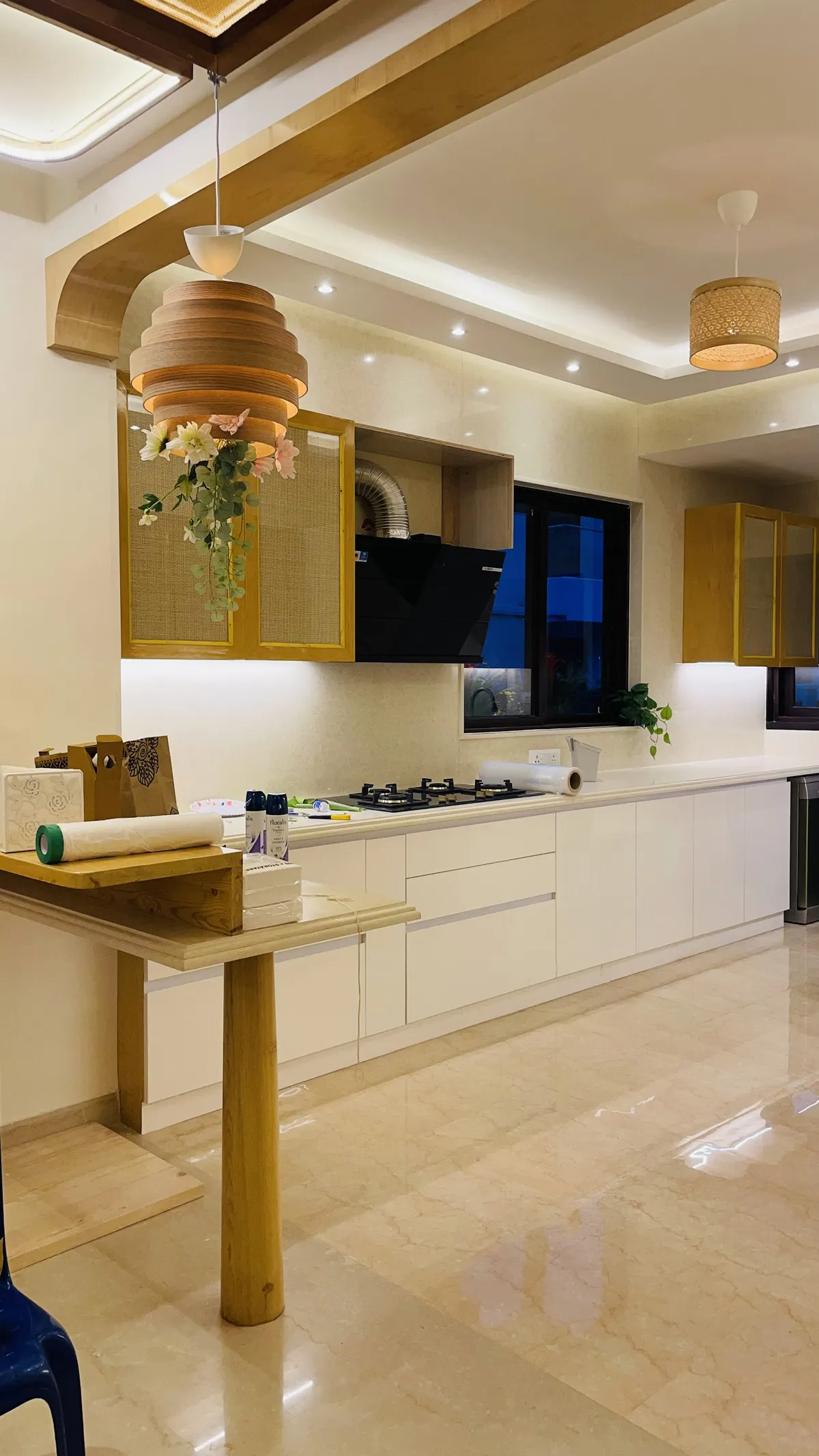 Long open kitchen shot at night: white handleless base units under a cream stone counter, cane-fronted wall cabinets in teak frames