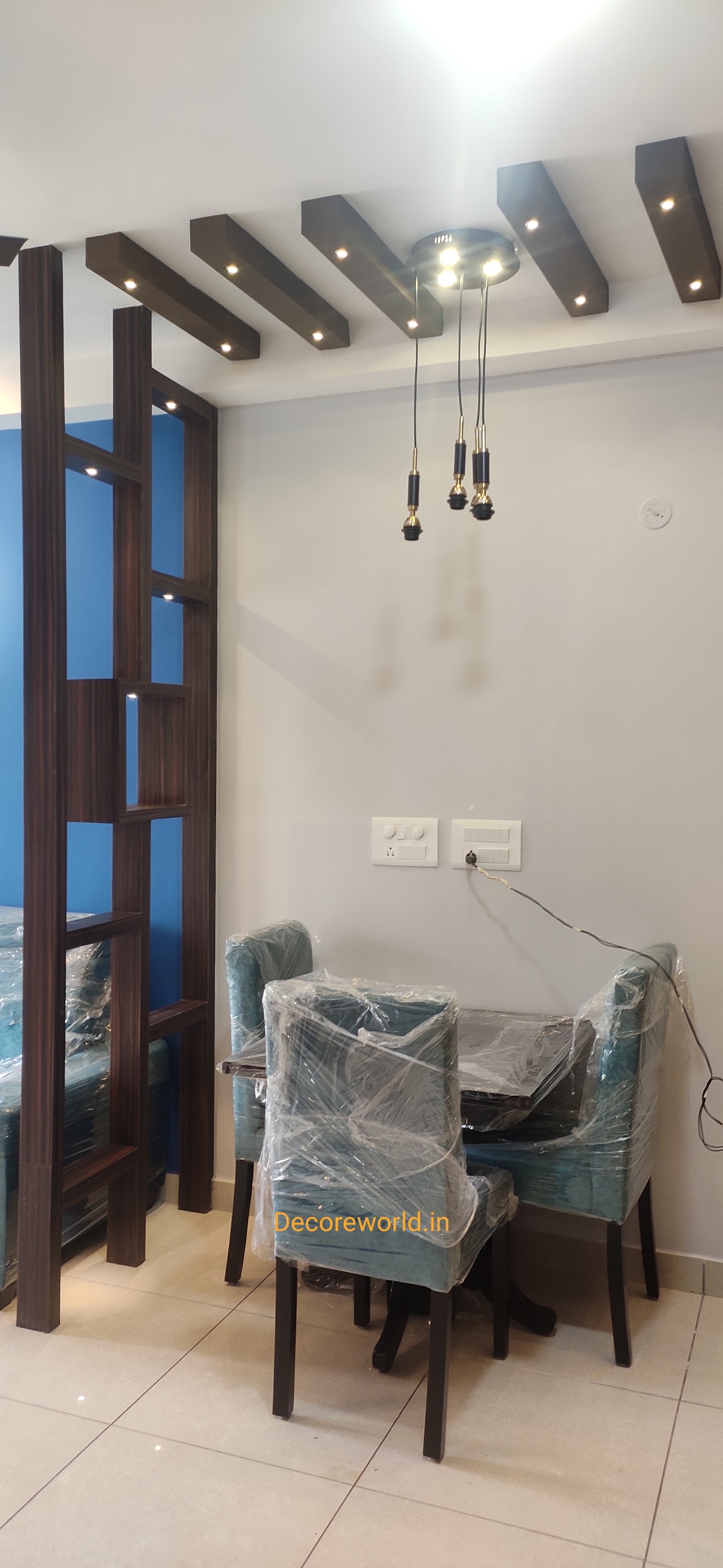 Newly finished dining nook with a wood-beam ceiling, brass-finished pendant lights and a blue accent wall, furniture still wrapped in protective plastic.
