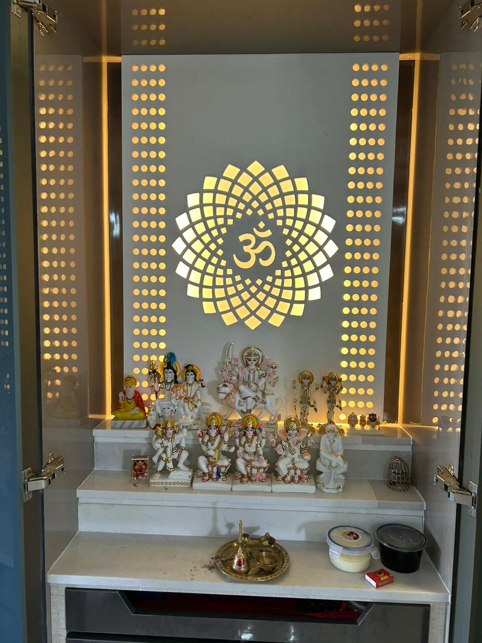 Pooja unit with a backlit lotus-and-Om medallion feature panel, perforated jaali side panels, and a tiered marble platform holding an array of deity idols.