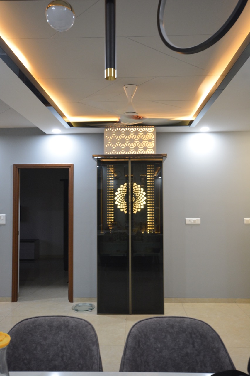 Dining area view of a floor-to-ceiling backlit black-glass pooja unit with a lotus-motif jaali screen, set beneath a stepped cove-lit false ceiling.