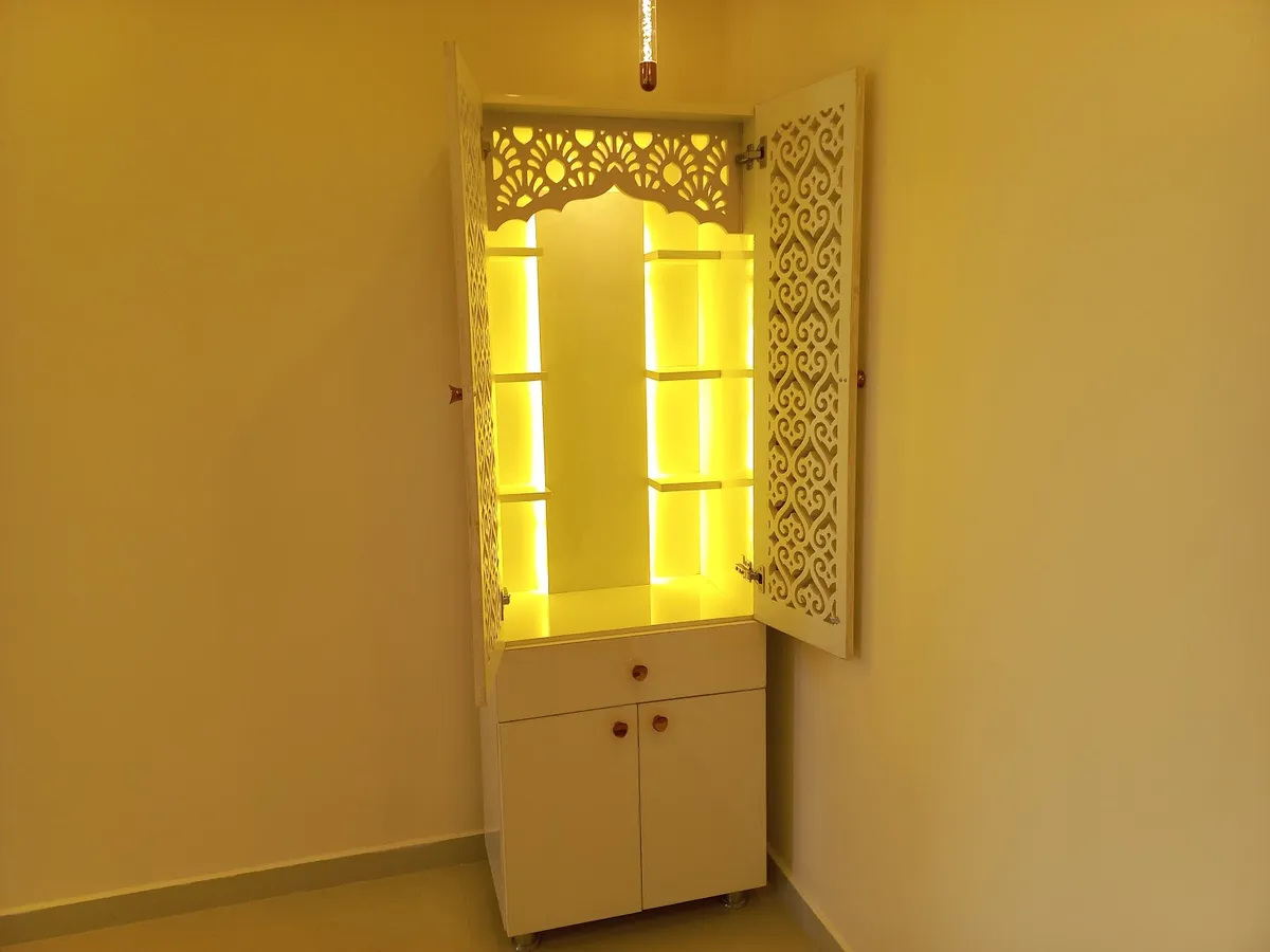 Freestanding white mandir with CNC jaali shutters swung open on a warm backlit niche of stepped shelves under a scalloped arch