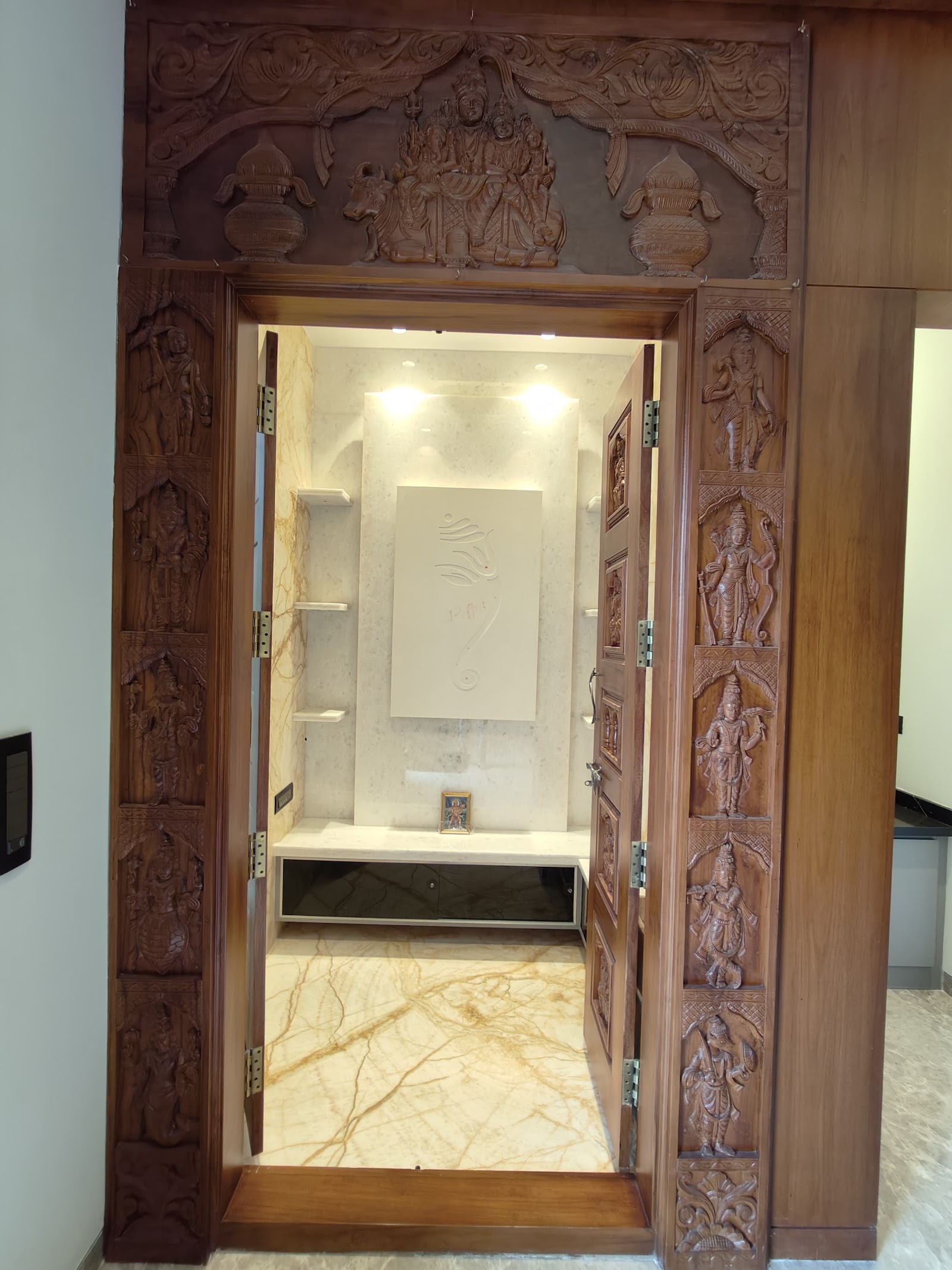 Pooja room entrance with an elaborately carved wood door depicting deities on the frame and panels