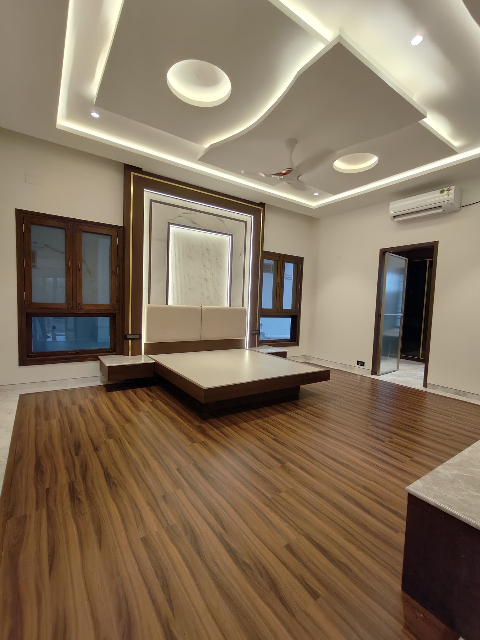 Master bedroom with a floating wood platform bed against a backlit marble headboard wall under a multi-level cove-lit ceiling