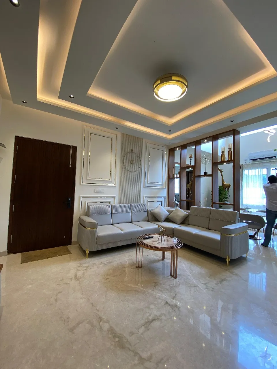 Living room with a pyramid-shaped gold-cove POP ceiling, moulded wall panelling flanking the main door, a wood-and-glass display partition