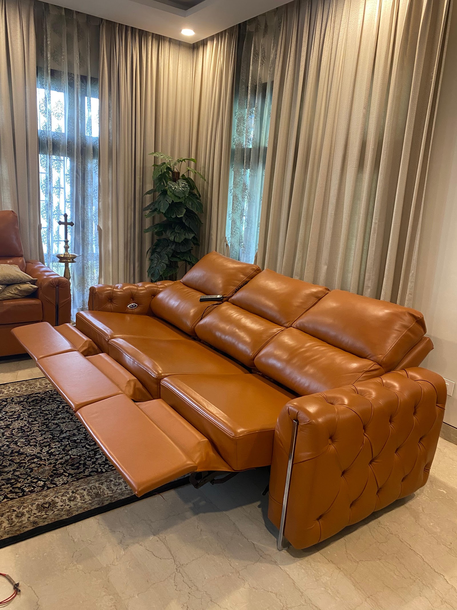 A tan tufted-leather reclining sofa shown fully extended, in a living room with floor-to-ceiling patterned sheer curtains, a traditional rug