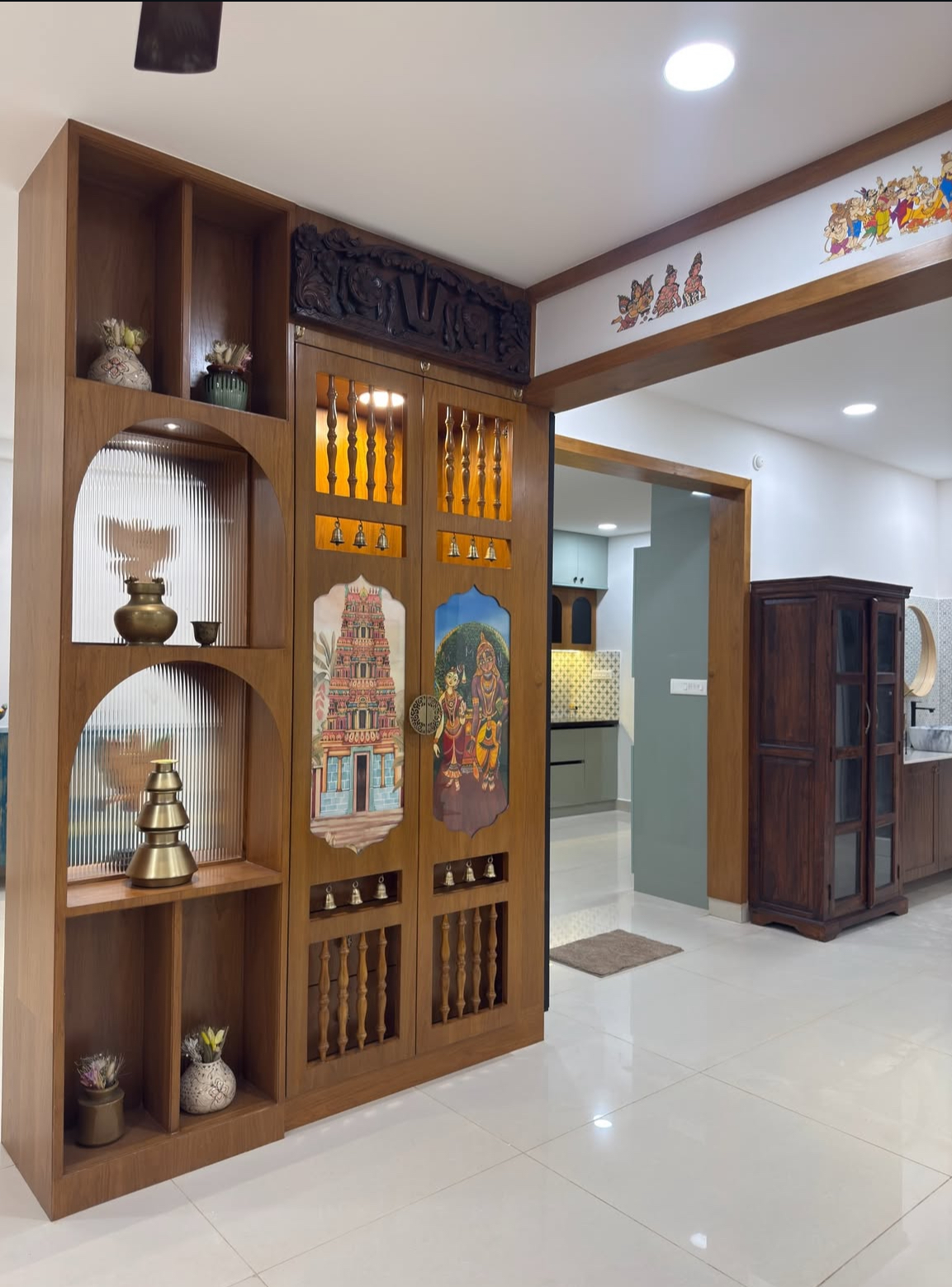 A pooja unit with a carved wood pediment, hand-painted temple and deity motifs on the cabinet doors, brass bells