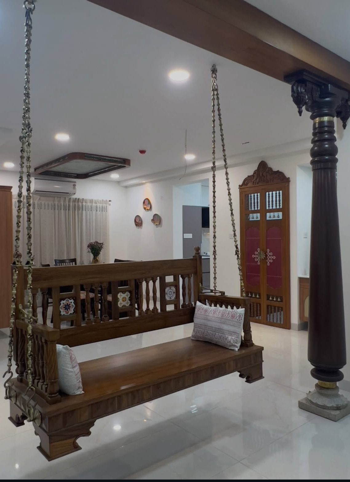 Wooden hanging swing on brass chains in an entrance hall, beside a carved wood pillar and a maroon-and-gold pooja door.