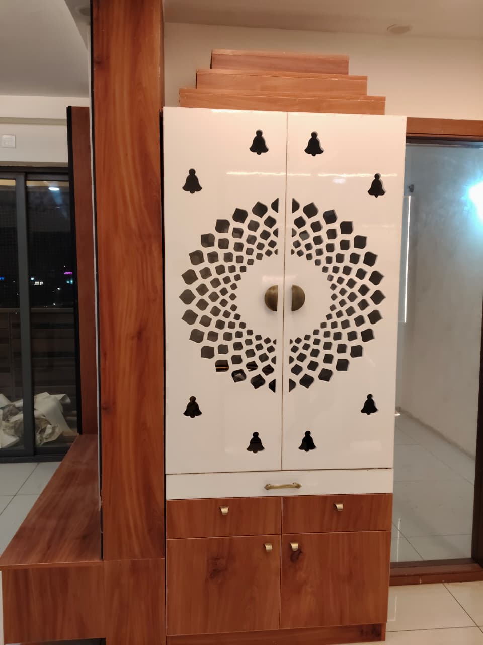 White high-gloss laminate pooja unit with laser-cut bell-motif jaali doors and wood-grain laminate drawer base, set against a foyer wall at night.