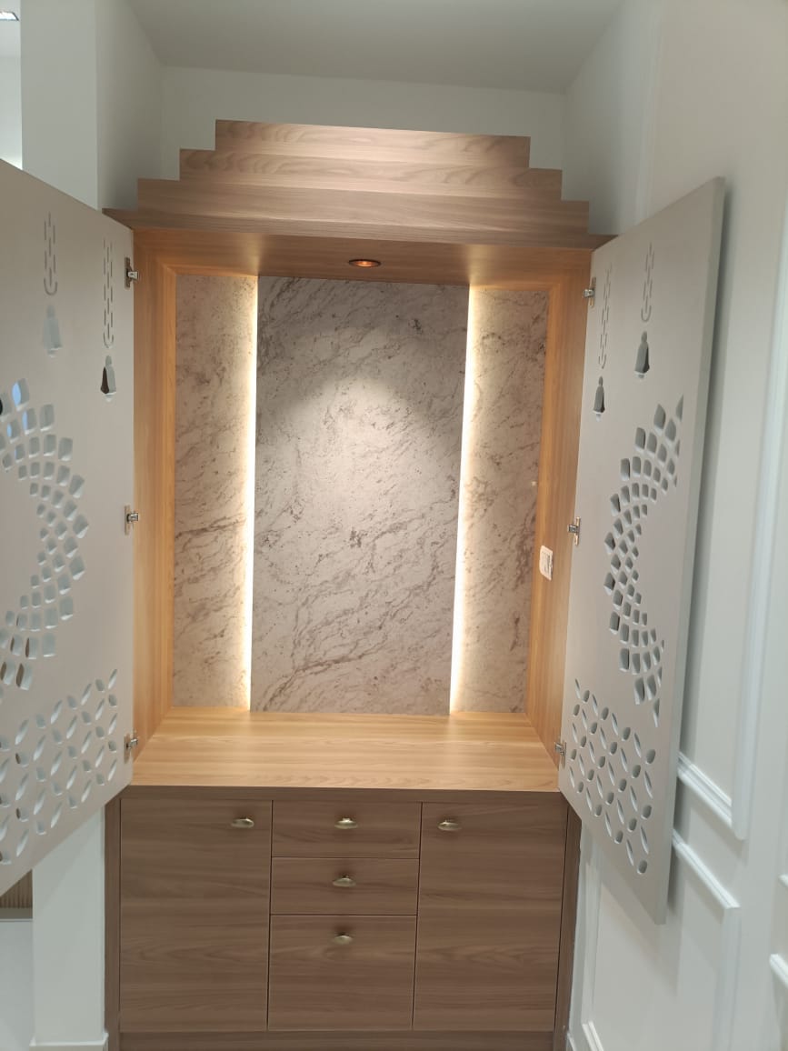 Pooja unit with open CNC-cut jaali doors revealing a backlit marble niche and tiered wood pediment above a walnut base cabinet with brass hardware.