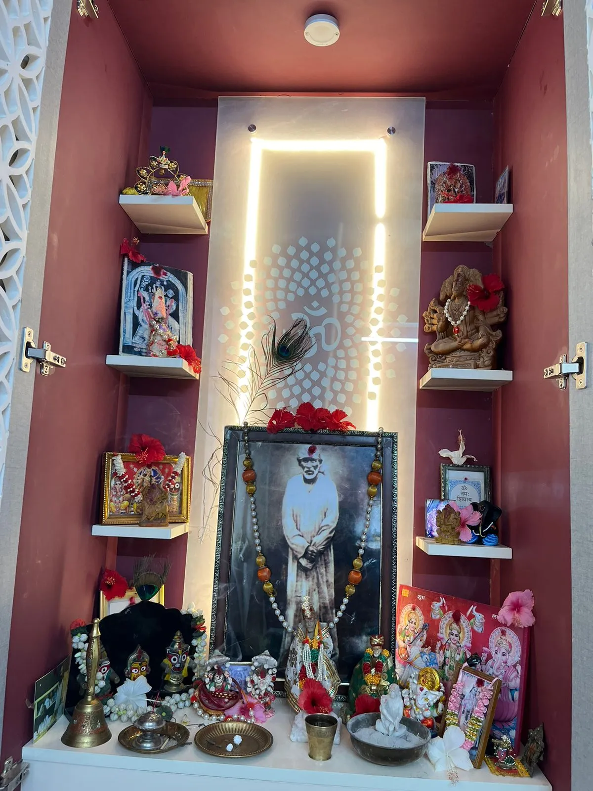 A home pooja unit interior with a backlit Om motif mirror panel, a framed photo of a saint with garlands