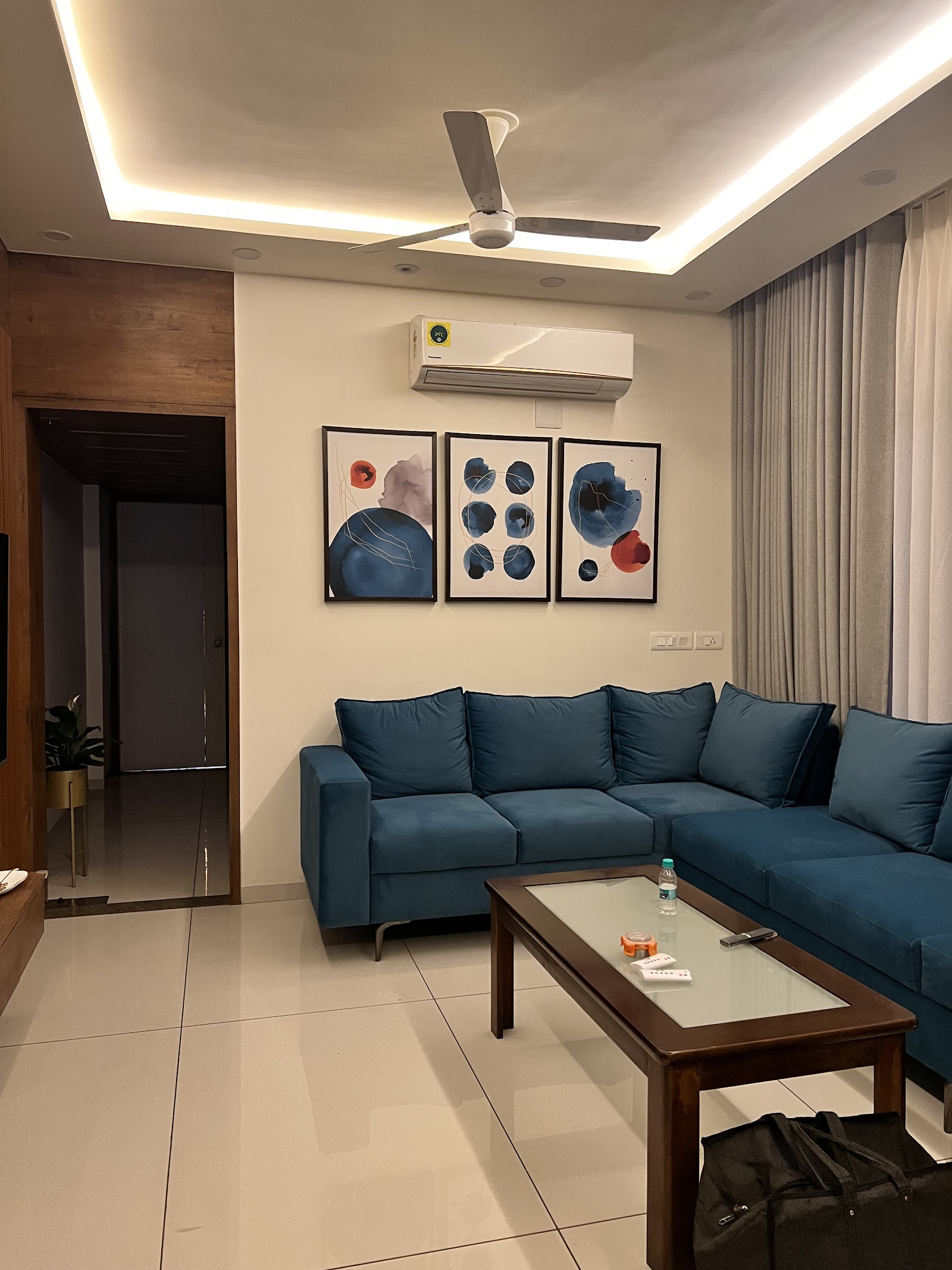 Living room with a deep teal L-shaped sofa, a three-panel abstract art gallery wall, a wood-and-glass coffee table, and a cove-lit ceiling.