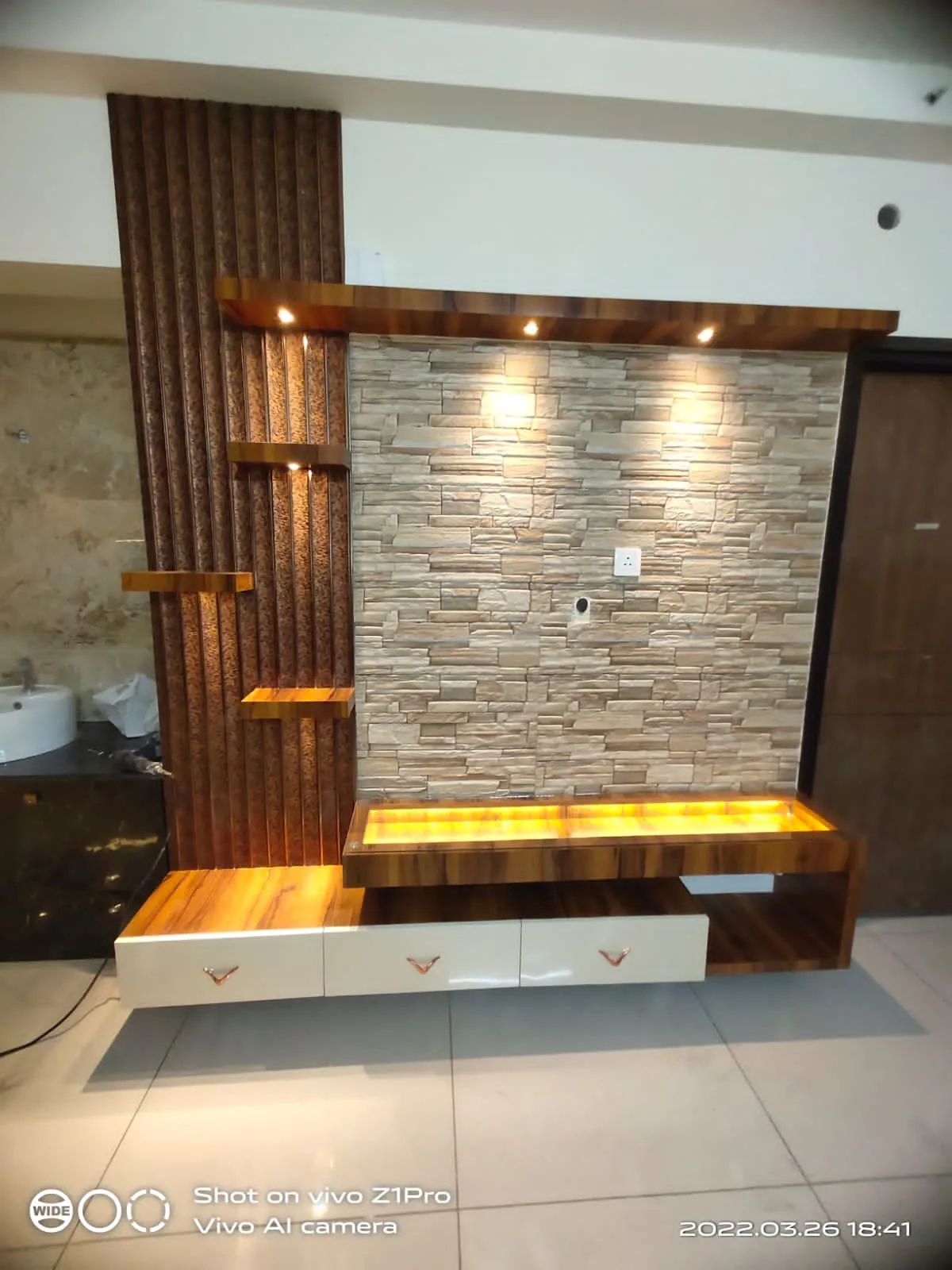 Living room TV feature wall combining a stacked-stone-look tile backdrop with a vertical fluted wood panel and a backlit wood floating unit with white