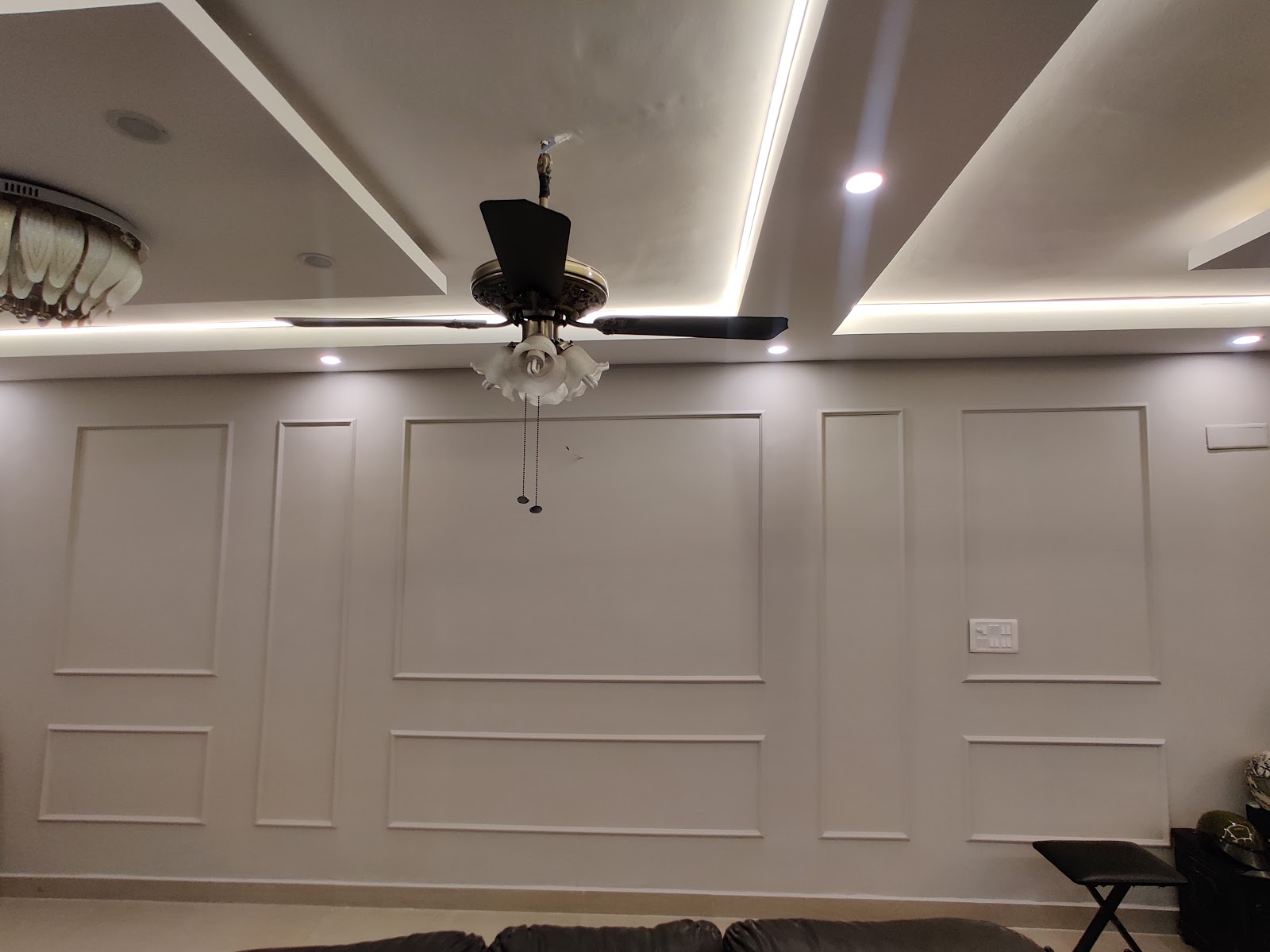 A living room accent wall with full-height panel moulding beneath a layered cove-lit false ceiling and an ornate ceiling fan with frosted glass shades.
