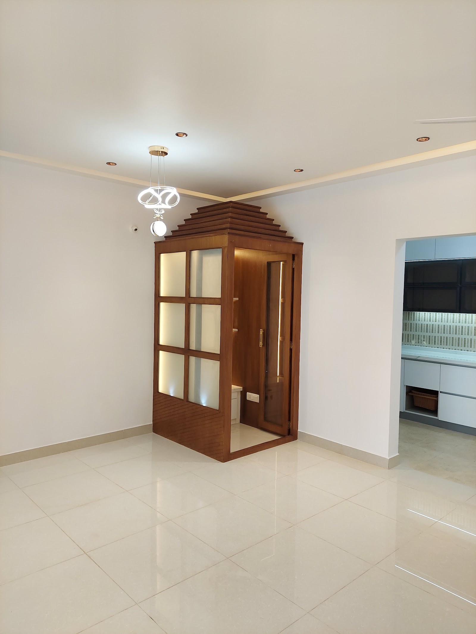 Freestanding wooden pooja unit with a tiered temple-style roof and frosted-glass shutters in the corner of an open-plan living area.