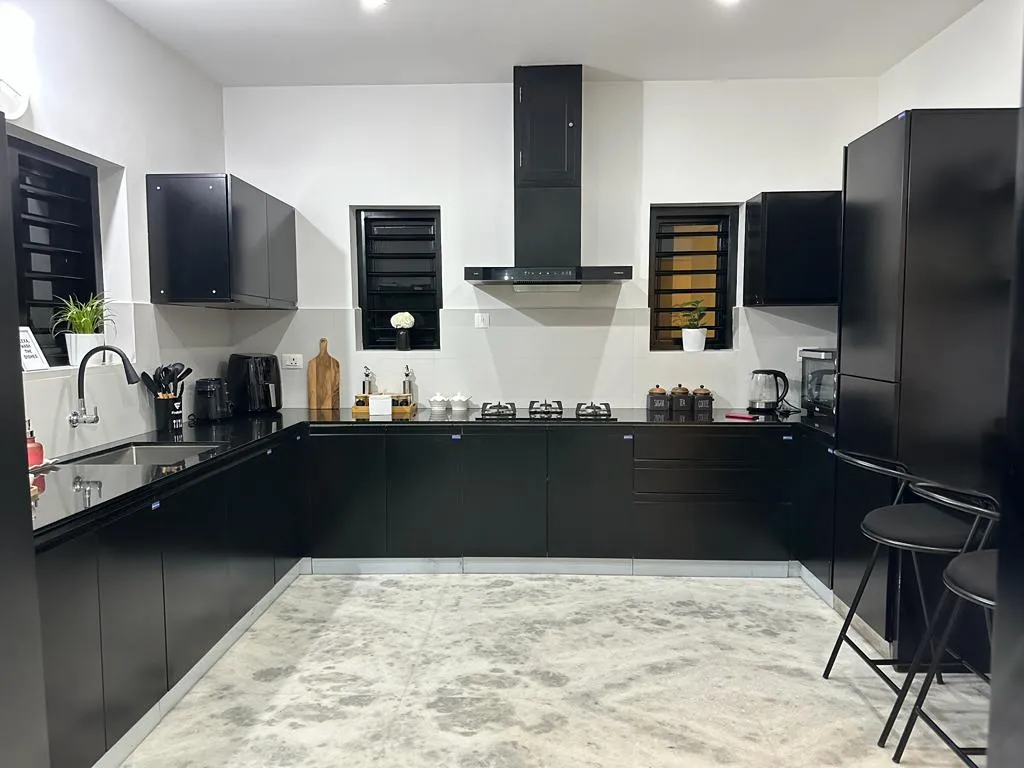 All-black U-shaped kitchen with matte black cabinetry, a black chimney hood, black granite countertops, black window grilles, and bar seating along one edge.