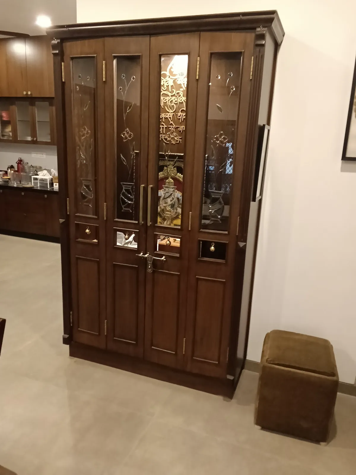 Tall teak mandir with etched-glass shutters carrying a gold shloka down the centre panel and a brass temple latch