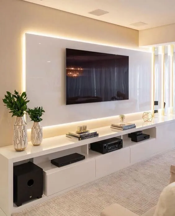 A living room TV wall with a glossy white backlit panel above a floating console styled with textured vases, stacked books, and a geometric-patterned rug.