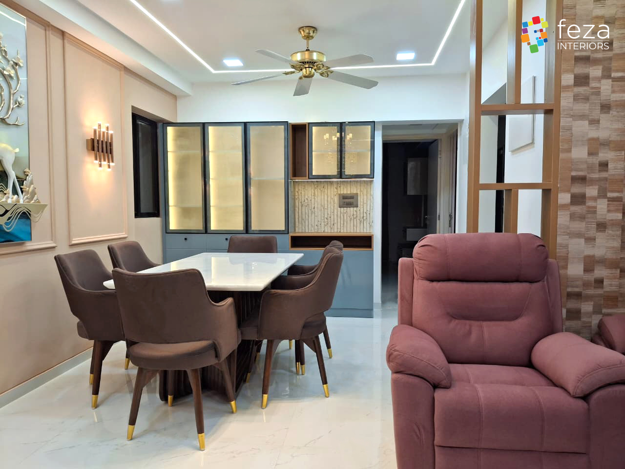Dining nook with a marble-top table, brass-capped velvet armchairs and a backlit glass crockery unit, opening to a living area with a mauve recliner.