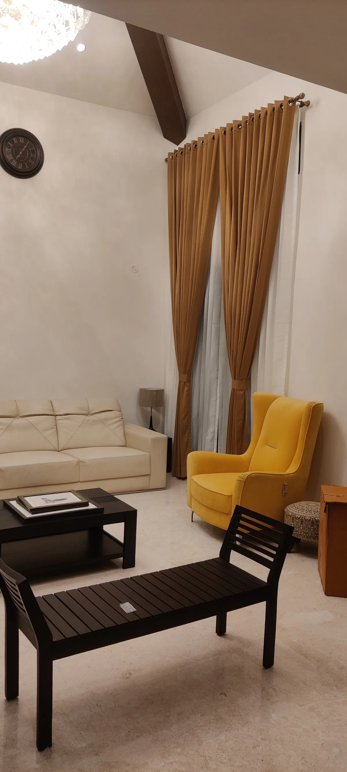 Sloping-ceiling living room with a dark exposed beam, cream leather three-seater, mustard velvet wingback and gold pinch-pleat curtains layered over white