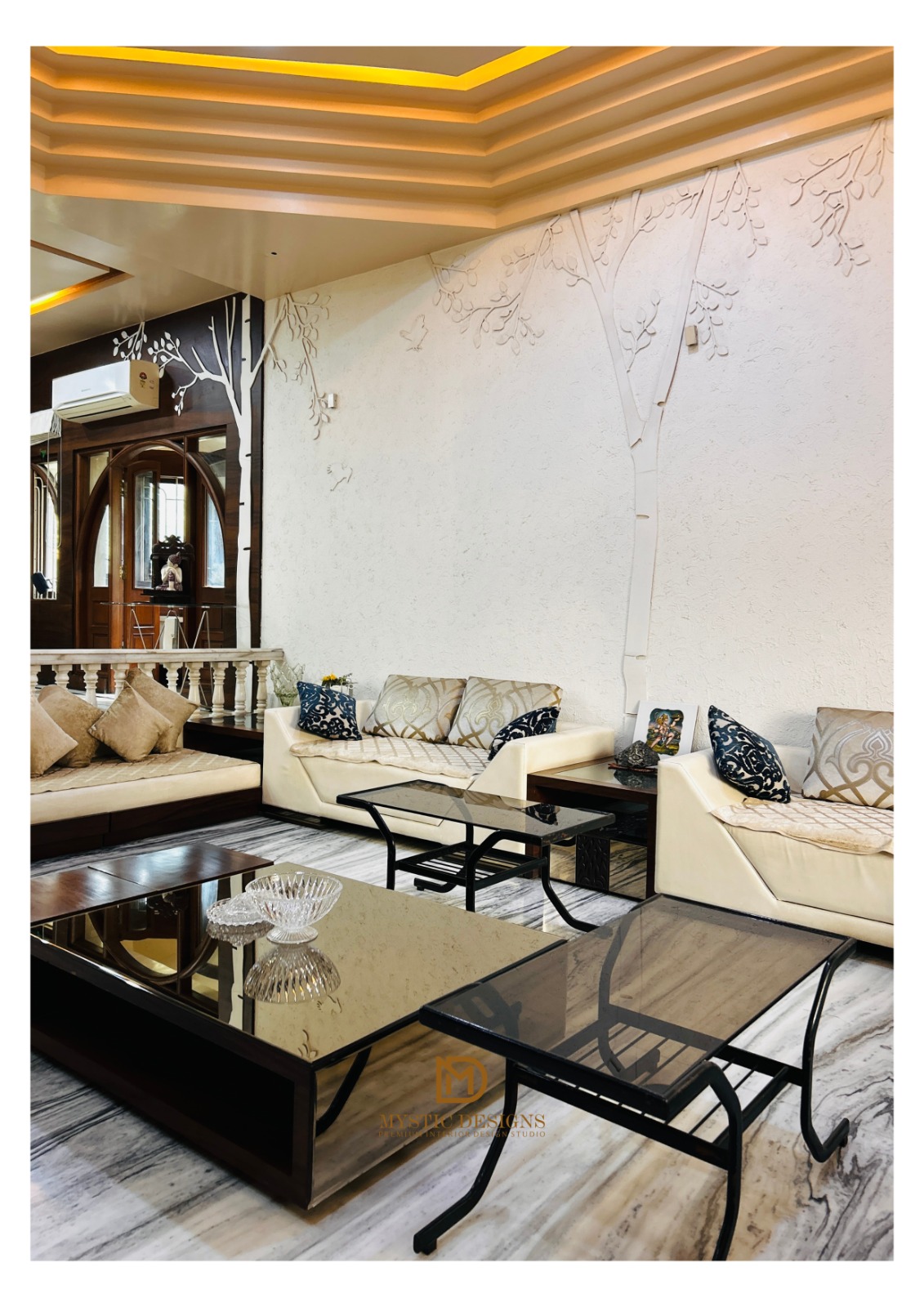 A living room with a 3D textured tree mural on the end wall, a gold cove-lit tray ceiling, cream leather sofas