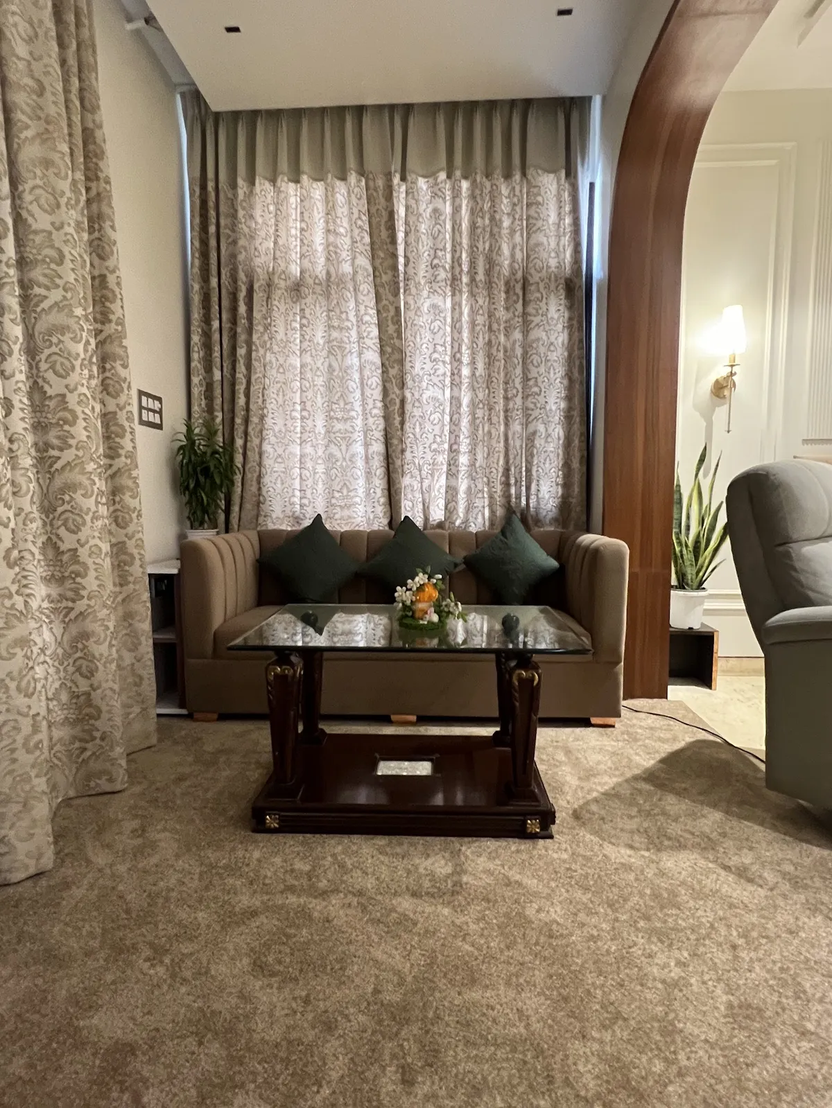 Living room with a carved dark wood and glass coffee table, deep green cushions on a beige sofa, and floor-to-ceiling damask curtains.