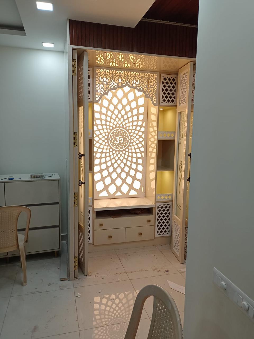 Backlit pooja unit with an intricately carved Om-motif jaali panel, arched niche, and folding CNC jaali doors flanking open shelves.