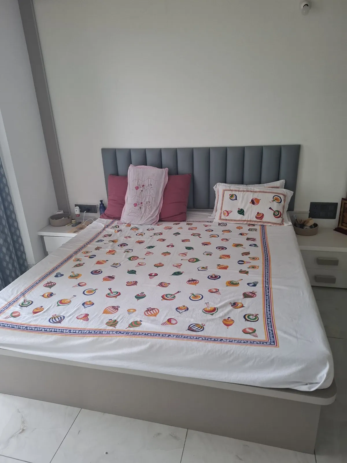 A grey channel-tufted floating bed with a colourful traditional block-print bedspread, flanked by white floating nightstands, against plain white walls.