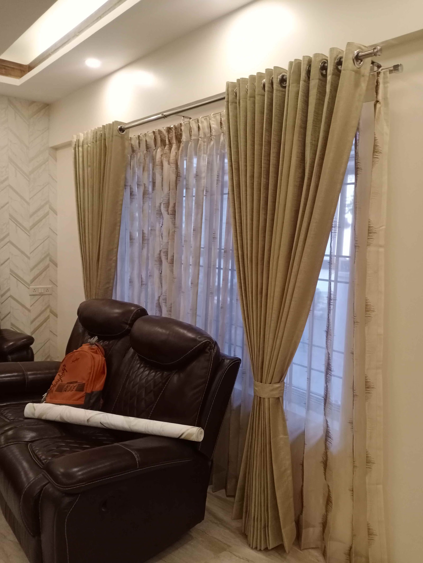 Living room corner with a black leather reclining sofa in front of layered gold curtains over sheer inner drapes, chevron-pattern wallpaper on the side wall.