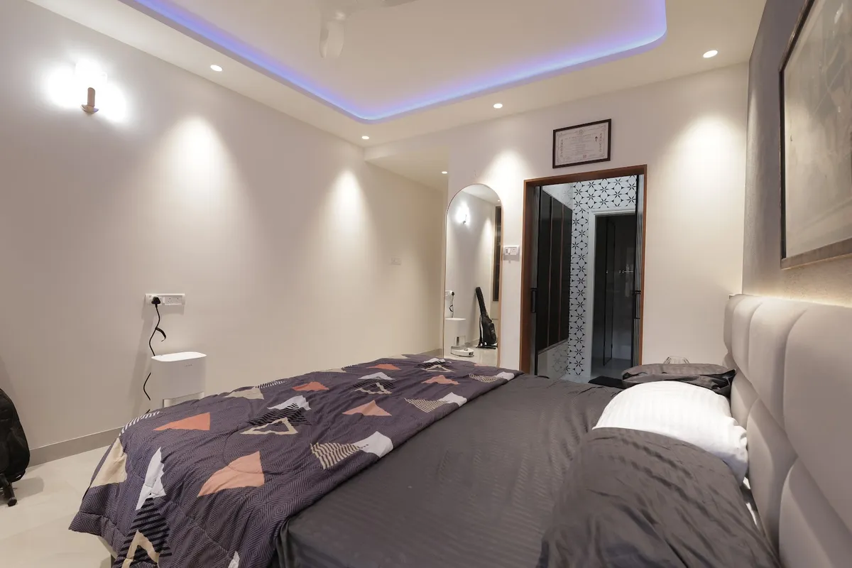 A grey-toned bedroom with a blue LED cove ceiling and a full-wall upholstered headboard, opening into an en-suite bath through a patterned glass door.