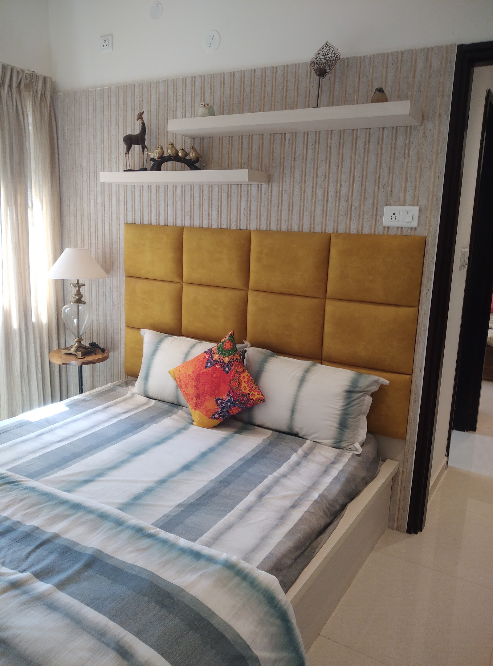 Master bedroom with a mustard-yellow tufted panel headboard against striped wallpaper, twin floating shelves styled with brass and wood curios