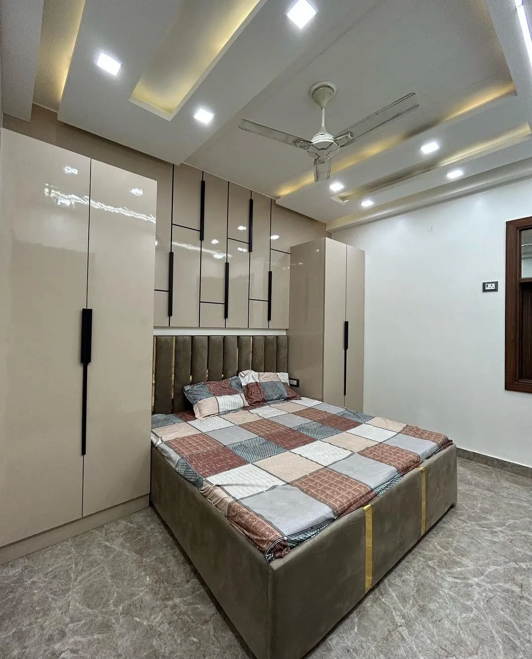 Real photo of a bedroom with a grey velvet channel-tufted headboard trimmed in gold, flanking beige glossy wardrobes with vertical black-strip inlays