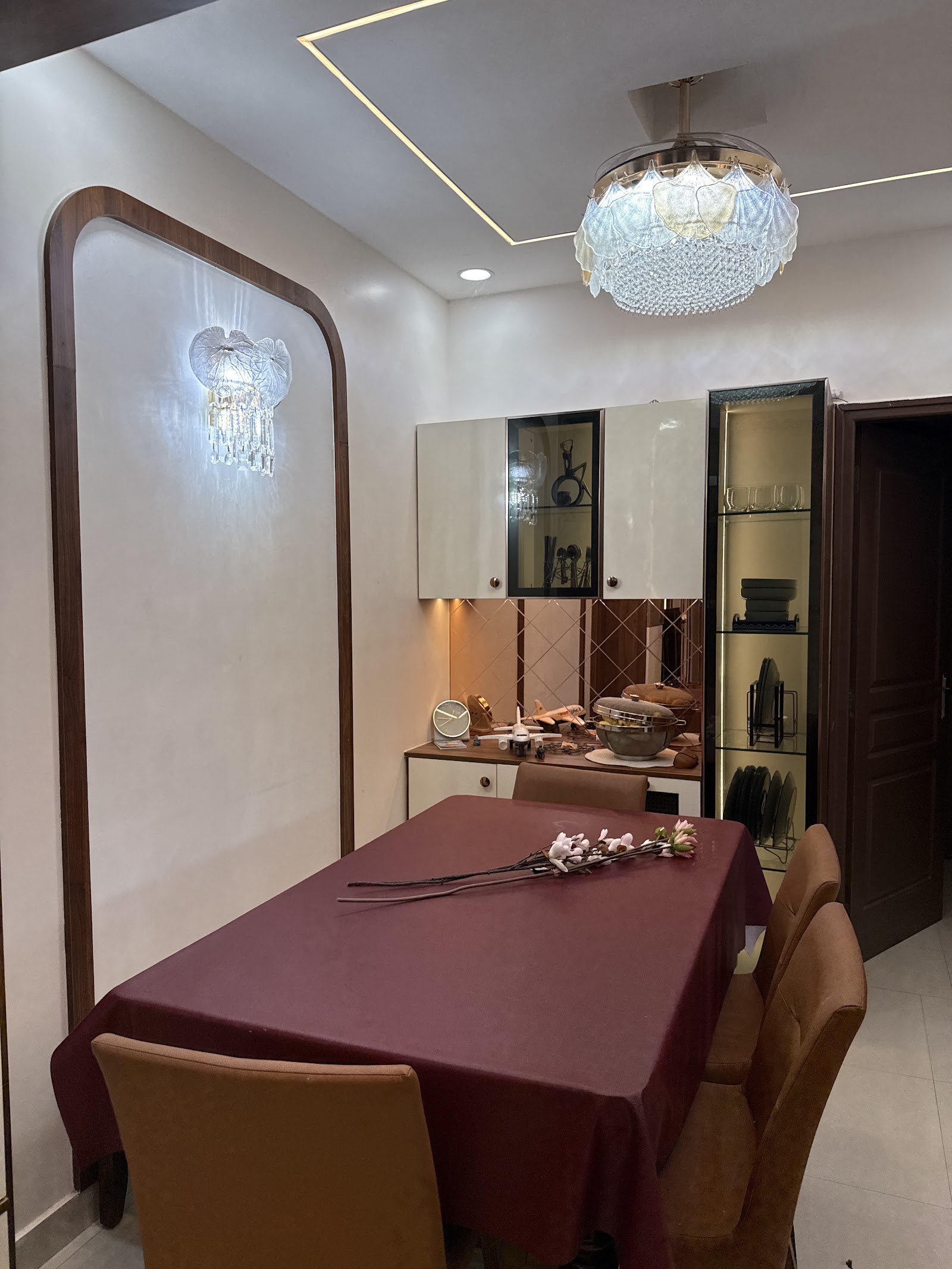 Dining room with a rectangular table under maroon linen, tan leather chairs, a crystal ceiling chandelier matched by a wall sconce set in an arched wood-framed