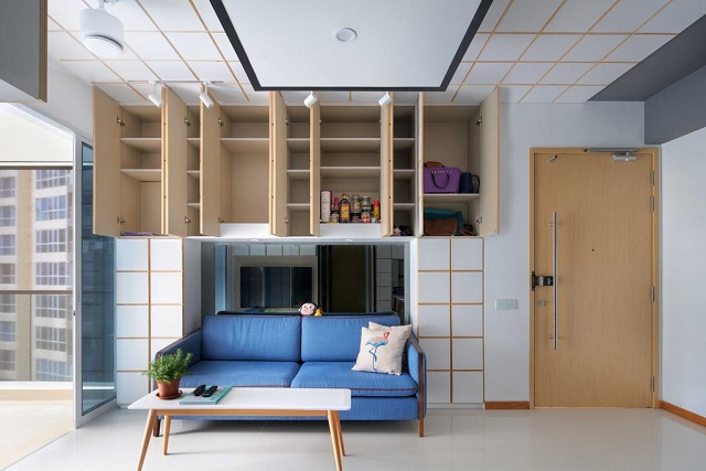 Compact living room with a blue sofa and white coffee table beneath open overhead storage cabinets in light wood, gridded false ceiling with track lighting