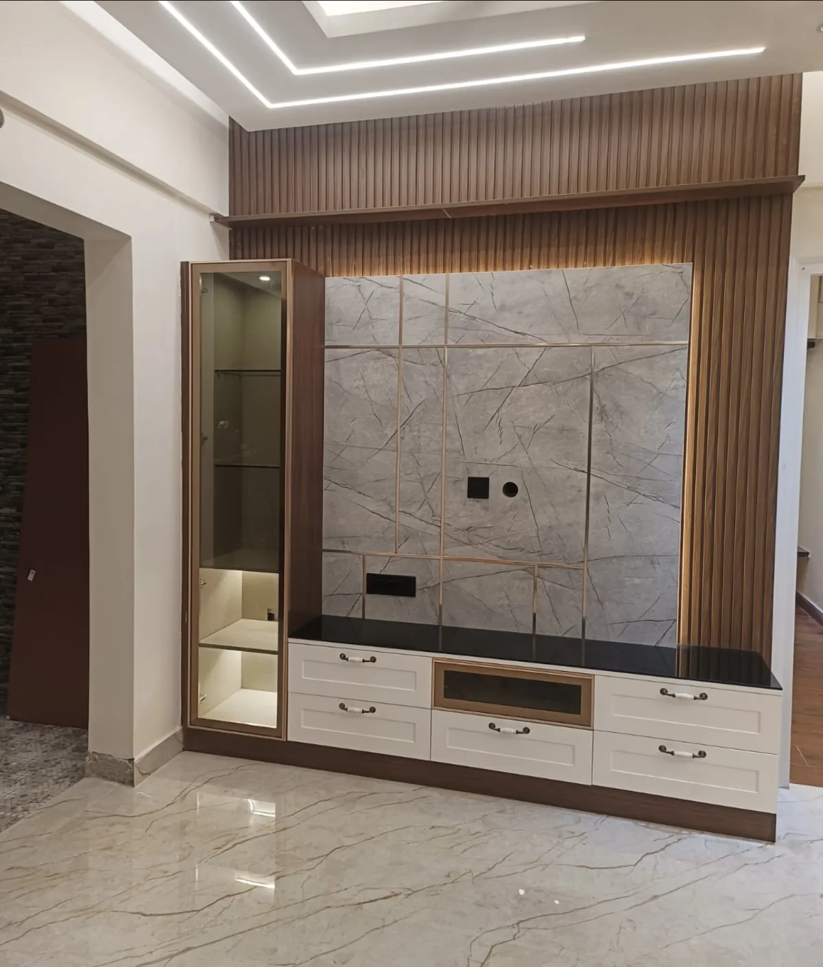 Real photo of a living room TV wall with fluted wood panelling framing a brass-inlaid grey marble slab, a glass-front display cabinet