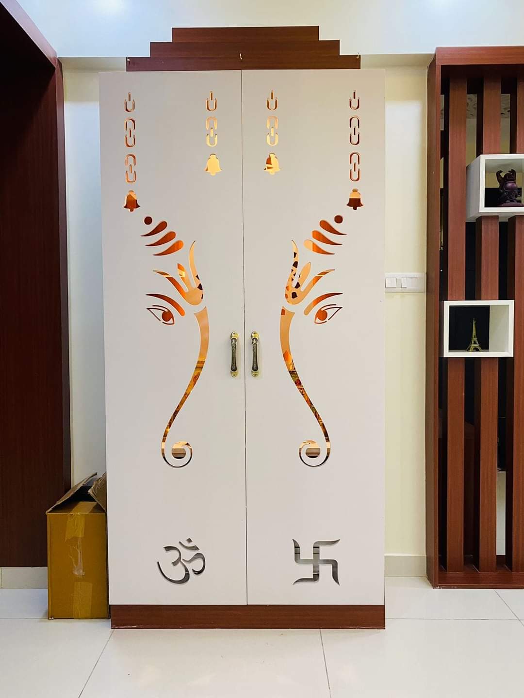 Free-standing white pooja cabinet with a mirrored CNC-cut Ganesha-ear motif and Om and swastik cutouts on the double doors, brass handles.