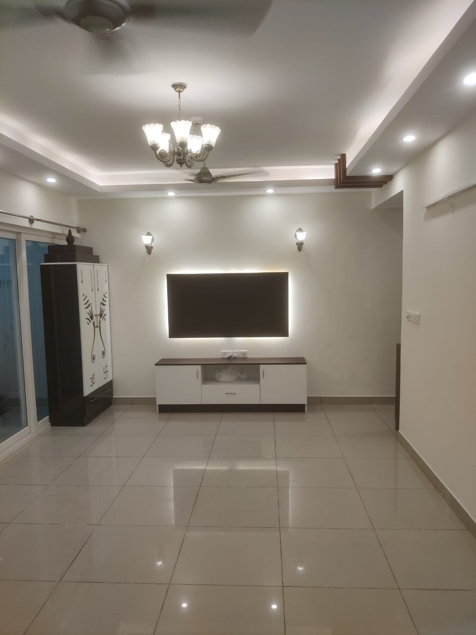 Living room with a backlit TV feature wall, a chandelier under a stepped false ceiling, and a tall pooja cabinet by the balcony door